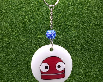 May include: A white, round keychain with a cartoon robot face in red and black. The keychain has a silver chain and ring, with a blue beaded accent. The background is a textured green surface.