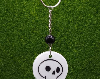 May include: A white, circular keychain pendant featuring a black skull design. The keychain includes a silver-colored key ring, a twisted chain, and a black bead. The background is a solid green color.