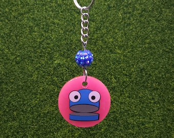 May include: A keychain featuring a pink circular charm with a cartoon character design in blue and gray. The charm is attached to a silver chain, a blue beaded ball, and a silver keyring. The background is green.