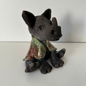 Może przedstawiać: Ceramiczna figurka nosorożca w zielonej, czerwonej i miedzianej kurtce. Figurka siedzi ze skrzyżowanymi nogami i ma szczegółową twarz.