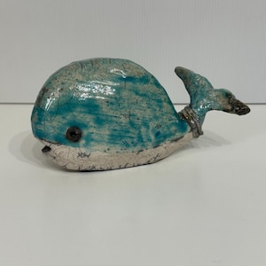 Puede incluir: Una escultura de ballena de cerámica azul turquesa con un glaseado blanco y un ojo negro. La ballena tiene una superficie texturizada y una pequeña aleta caudal.