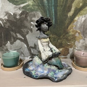 May include: Ceramic figurine of a woman with dark hair and a white dress sitting on a blue and green patterned base.