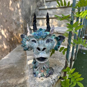Peut inclure: Une sculpture de tête de dragon en céramique détaillée avec une finition bleu-vert mouchetée texturée. Le dragon présente des cornes noires, des accents bleus et verts et des traits faciaux détaillés. La sculpture est exposée à l'extérieur.