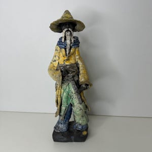 Puede incluir: Figura de cerámica de un mago con abrigo amarillo, pantalones verdes y azules y sombrero de ala ancha. El mago tiene una larga barba blanca y está sobre una base oscura. La figura tiene un aspecto texturizado y envejecido.
