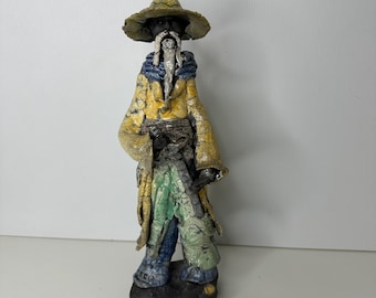 Puede incluir: Figura de cerámica de un mago con abrigo amarillo, pantalones verdes y azules y sombrero de ala ancha. El mago tiene una larga barba blanca y está sobre una base oscura. La figura tiene un aspecto texturizado y envejecido.