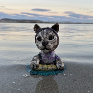 May include: A ceramic cat figurine wearing a purple and green swimsuit, floating on a yellow and blue inner tube. The cat has a white and gray patterned body and is set against a blurred background of a beach and water.