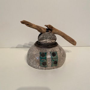 Op de afbeelding: Een witte keramische pot met een gekrakelde glazuur en een stuk drijfhout dat aan de bovenkant is bevestigd. De pot heeft vier vierkante accenten met een tealblauw en zwart ontwerp.