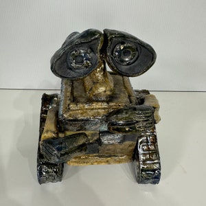 Może przedstawiać: Ceramiczna rzeźba robota z prostokątnym korpusem, dużymi czarnymi oczami i fakturowanym wykończeniem w kolorze brązowym i czarnym.