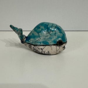 Op de afbeelding: Een klein keramiek walvisbeeldje met een blauwe en witte glazuur. De walvis heeft een gekrakelde glazuureffect, waardoor het een unieke en rustieke uitstraling heeft.