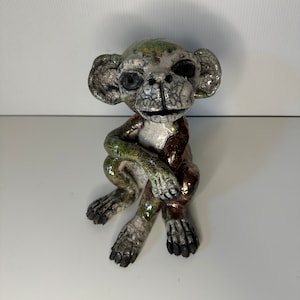 Może przedstawiać: Ceramiczna figurka małpy w zielonym, brązowym i białym szkliwie. Małpa siedzi z założonymi rękami i ma przebiegły wyraz twarzy.