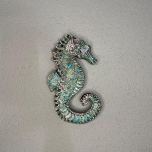 Op de afbeelding: Een gedetailleerde zeepaardbroche met een zilverkleurige afwerking. Het zeepaardje heeft ingewikkelde details met turquoise, roze en groene emaille accenten. De broche heeft een gebogen staart en een gedetailleerde kop.