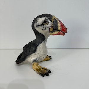 Peut inclure: Une sculpture en céramique d'un macareux à plumes noires et blanches, un bec rouge et jaune et des pattes jaunes. Le macareux est debout sur une surface blanche.