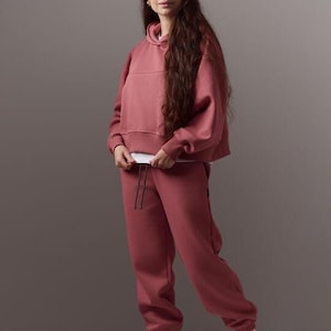 May include: A matching mauve-colored hoodie and sweatpants set. The hoodie has a drawstring and a cropped fit. The sweatpants have a relaxed fit with a drawstring waist. The outfit is paired with white high-top sneakers.