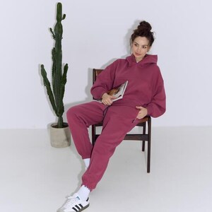 Pode incluir: Um conjunto de moletom com capuz e calças de moletom bordô combinando. O moletom com capuz tem capuz com cordão e um bolso frontal. As calças de moletom têm cintura e punhos elásticos. A pessoa está usando meias brancas e tênis Adidas com listras pretas.