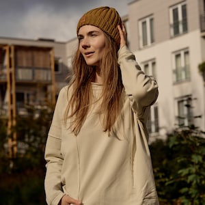 May include: A woman wearing a beige sweatshirt, blue jeans, and a brown beanie. She is standing outdoors in front of a building.