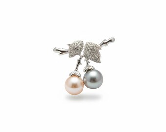 May include: A silver-toned brooch shaped like a cherry branch with two leaves encrusted with small clear stones. The brooch features two dangling spherical pearls, one in a soft pale pink hue and the other in a deep metallic gray color.
