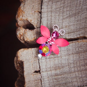 Puede incluir: Un broche de mariposa de color rosa y morado con detalles plateados y adornado con piedras preciosas. El broche está sobre una pieza de madera desgastada.