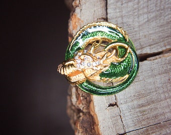 Puede incluir: Un broche de dragón de esmalte dorado y verde con diamantes de imitación. El broche está sobre una superficie de madera.