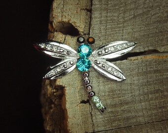 May include: A silver dragonfly brooch with blue and green rhinestones. The brooch is set against a rustic wooden background.