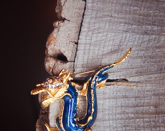 Puede incluir: Un broche de dragón de esmalte azul y dorado con diamantes de imitación. El dragón está enroscado con la cola en la boca.