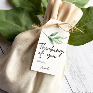 May include: A small, beige fabric bag with a handwritten tag that reads "Thinking of you" with a green leafy design. The tag also includes the text "With love, Amanda".