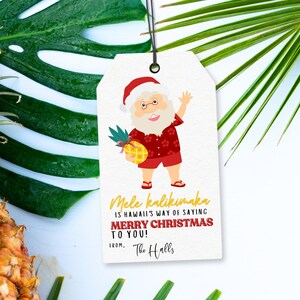 May include: A white gift tag with a cartoon illustration of Santa Claus wearing a red and white Santa hat, red shorts, and a red and white Hawaiian shirt. He is holding a pineapple. The text on the tag reads "Mele Kalikimaka is Hawaii's way of saying Merry Christmas to you! From, The Halls."