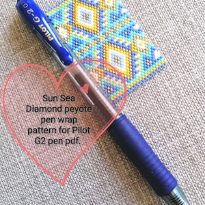 May include: A blue pen with the text "PILOT G-2 07" on the side, lying on a beige fabric surface. A small beaded square with a geometric pattern in shades of blue, yellow, and white is partially visible in the background. The text "Sun Sea Diamond peyote pen wrap pattern for Pilot G2 pen pdf." is written in a pink heart shape.