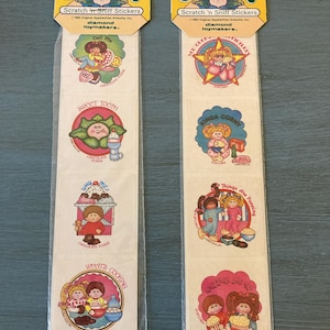 May include: Two packages of Cabbage Patch Kids Scratch 'n Sniff Stickers. Each package has four stickers with different images and scents. The stickers feature Cabbage Patch Kids dolls in various scenes, including a doll on a phone, a doll with a sundae, a doll with a popcorn bucket, and a doll with a bowl of popcorn.