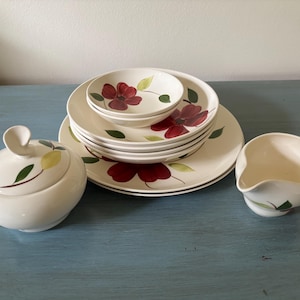 May include: A ceramic dinnerware set featuring a cream-colored base with a painted pattern of dark red flowers and green leaves. The set includes a lidded sugar bowl, a creamer, and a stack of plates and bowls of varying sizes.