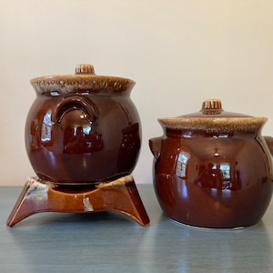 May include: Two brown glazed ceramic casserole dishes with lids. The smaller dish is on a brown glazed ceramic stand with three legs.
