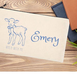 May include: A cream-colored zippered pouch featuring a blue illustration of a lamb with a bow, the name "Emery", and the text "WALK WITH ME". The pouch is on a wooden surface with books and a plant.