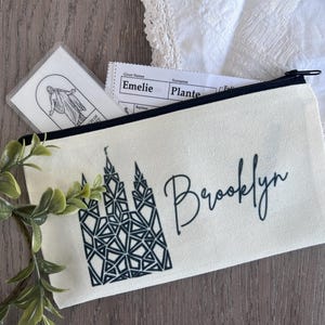 May include: A white canvas zippered pouch with a black and white illustration of a temple and the name "Brooklyn" in black script.