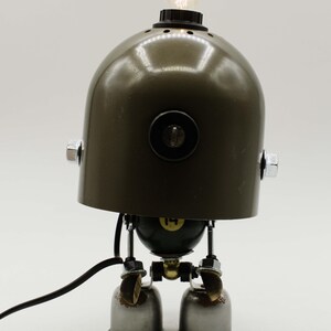 Puede incluir: Lámpara robot artesanal con una cabeza en forma de casco verde oliva oscuro y una pequeña bombilla. El cuerpo está hecho de piezas metálicas recicladas, incluyendo pequeños quemadores para los pies. La lámpara tiene un cable de alimentación negro.