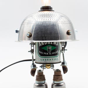May include: A silver robot lamp made from recycled materials. The robot has a colander for a head, a can for a body, and two metal cans for legs. The can has the text "Andrews Sali per il fegato Lassativi" on it.