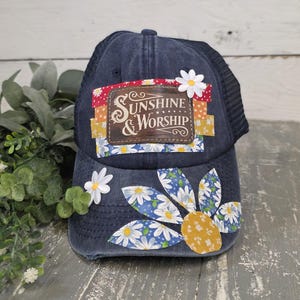May include: A navy blue trucker hat with a mesh back and a curved brim. The front features a patch with the words "Sunshine & Worship" and a daisy design. Additional daisy accents are placed on the hat.