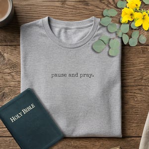 May include: A gray t-shirt with the text "pause and pray" is displayed on a wooden surface, alongside a dark blue Holy Bible and a cup of coffee. Yellow flowers and greenery add a touch of color.