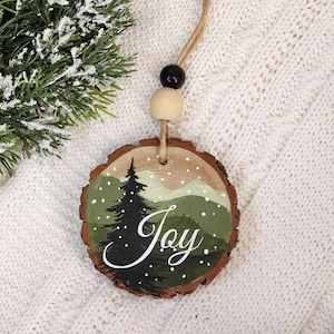 May include: A round wooden Christmas ornament with a painted winter scene. The design features a dark green pine tree, rolling green hills, and falling white snow. The word "Joy" is written in white script. The ornament is suspended by a twine loop with black and natural wooden beads.