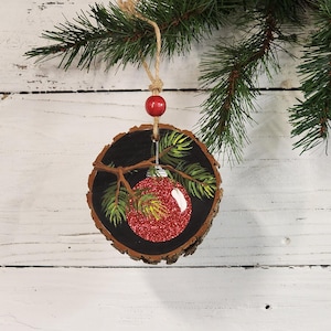 May include: A round, wooden Christmas ornament with a painted red glitter ball and green pine needles. The ornament hangs from a twine string and a red bead. The background is a white, wooden surface with green pine branches.