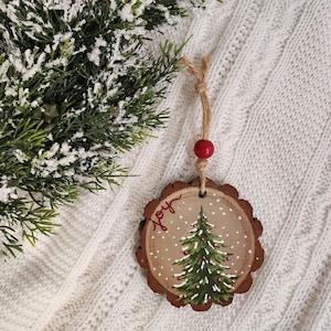 May include: A wooden Christmas ornament with a hand-painted green Christmas tree and the word "joy" in red. The ornament is round with a natural wood edge and a twine hanger. A red bead is attached to the twine. The background is a white knitted blanket.