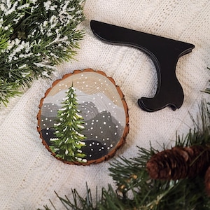 May include: A hand-painted wooden ornament featuring a winter landscape with a green pine tree and snowy mountains. The ornament is round and sits next to a black stand. Decorative evergreen branches and pine cones are also present.