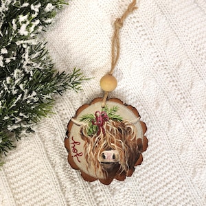 May include: A round wooden ornament featuring a hand-painted Highland cow with long, shaggy brown fur and a pink nose. The ornament has a natural wood grain edge and a twine hanger with a wooden bead. The word "joy" is written in red.