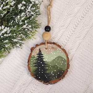 May include: A round, wooden Christmas ornament with a hand-painted winter scene. The design features a dark green pine tree, green hills, and white snowfall. The ornament is suspended by a natural twine cord with a black and wooden bead.