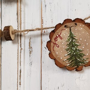 May include: A rustic wooden Christmas ornament with a painted green pine tree and the word "joy" in red script. The ornament is decorated with white paint to resemble snow.