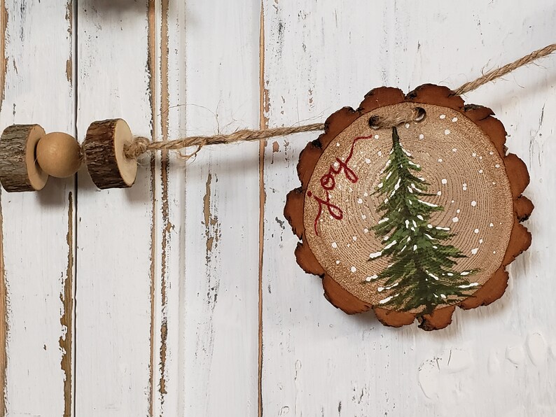 May include: A rustic wooden Christmas ornament with a painted green pine tree and the word "joy" in red script. The ornament is decorated with white paint to resemble snow.