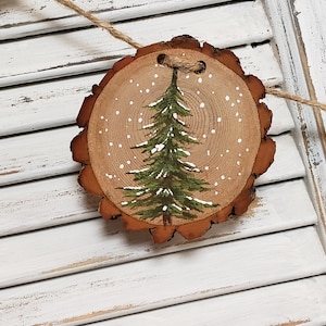 May include: A wooden Christmas ornament with a painted green pine tree and white snowflakes on a natural wood slice background.
