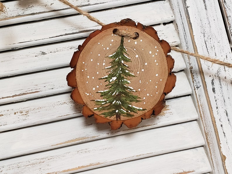 May include: A wooden Christmas ornament with a painted green pine tree and white snowflakes on a natural wood slice background.