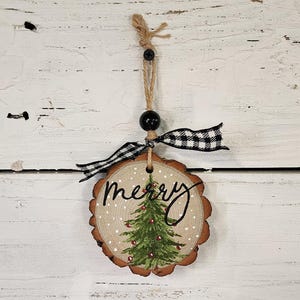 May include: A wooden Christmas ornament with a painted green Christmas tree and the word "merry" in black script. The ornament is round with a natural wood grain edge, a black and white checkered ribbon, and a jute string for hanging.