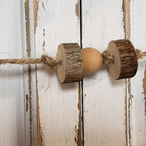 May include: A rustic wooden bead garland with a natural wood bead and twine string. The garland is hanging on a white painted wooden surface.
