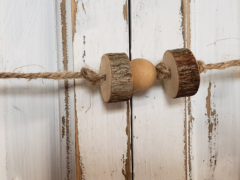 May include: A rustic wooden bead garland with a natural wood bead and twine string. The garland is hanging on a white painted wooden surface.