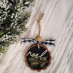 May include: A wooden Christmas ornament with a black painted surface and the word "noel" in white script. The ornament features a painted pine branch and is adorned with a black and white checkered ribbon and a wooden bead.